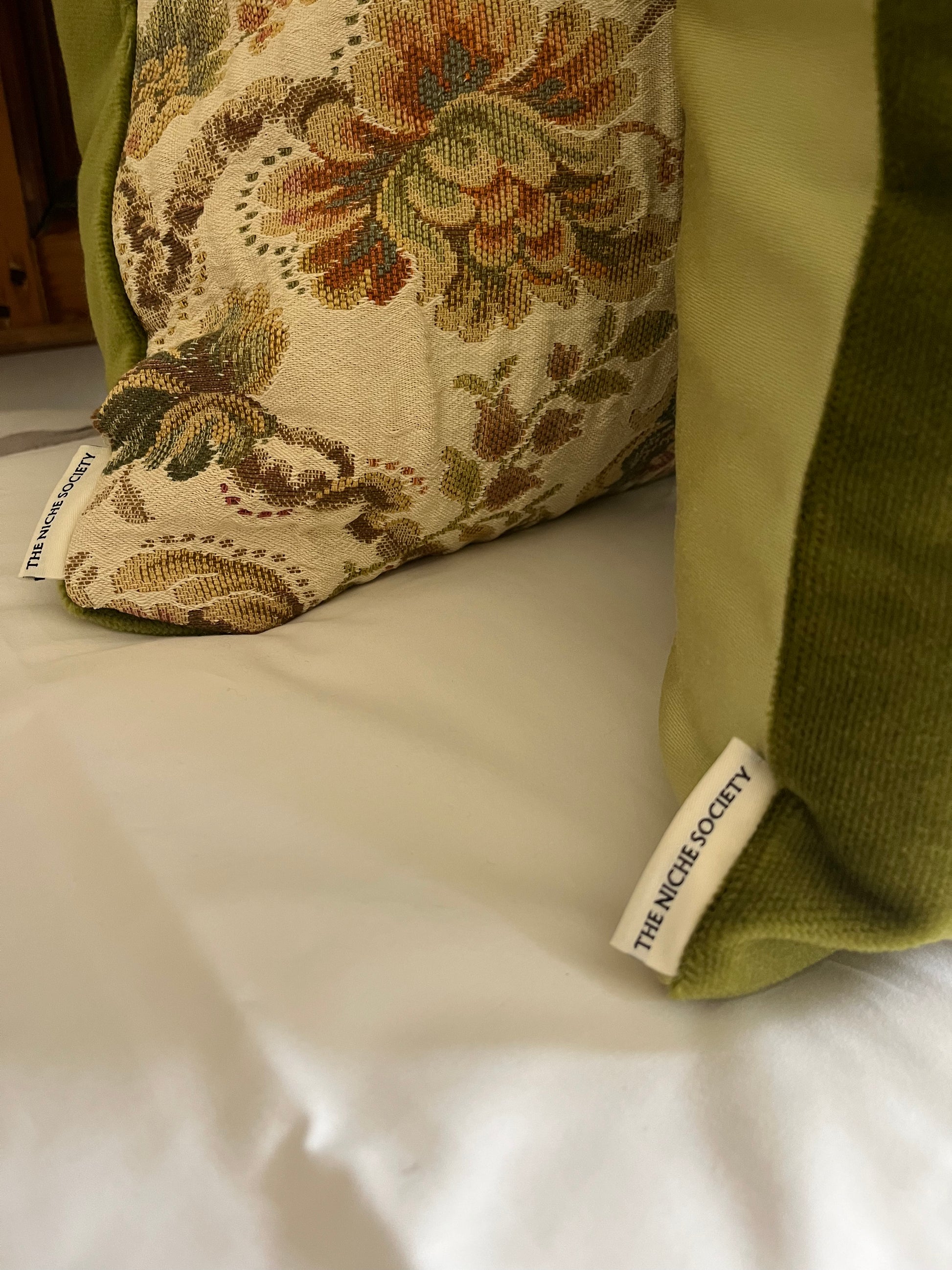 Decorative pillow with floral pattern and green velvet on a bed, featuring 'The Nicée Society' tag.